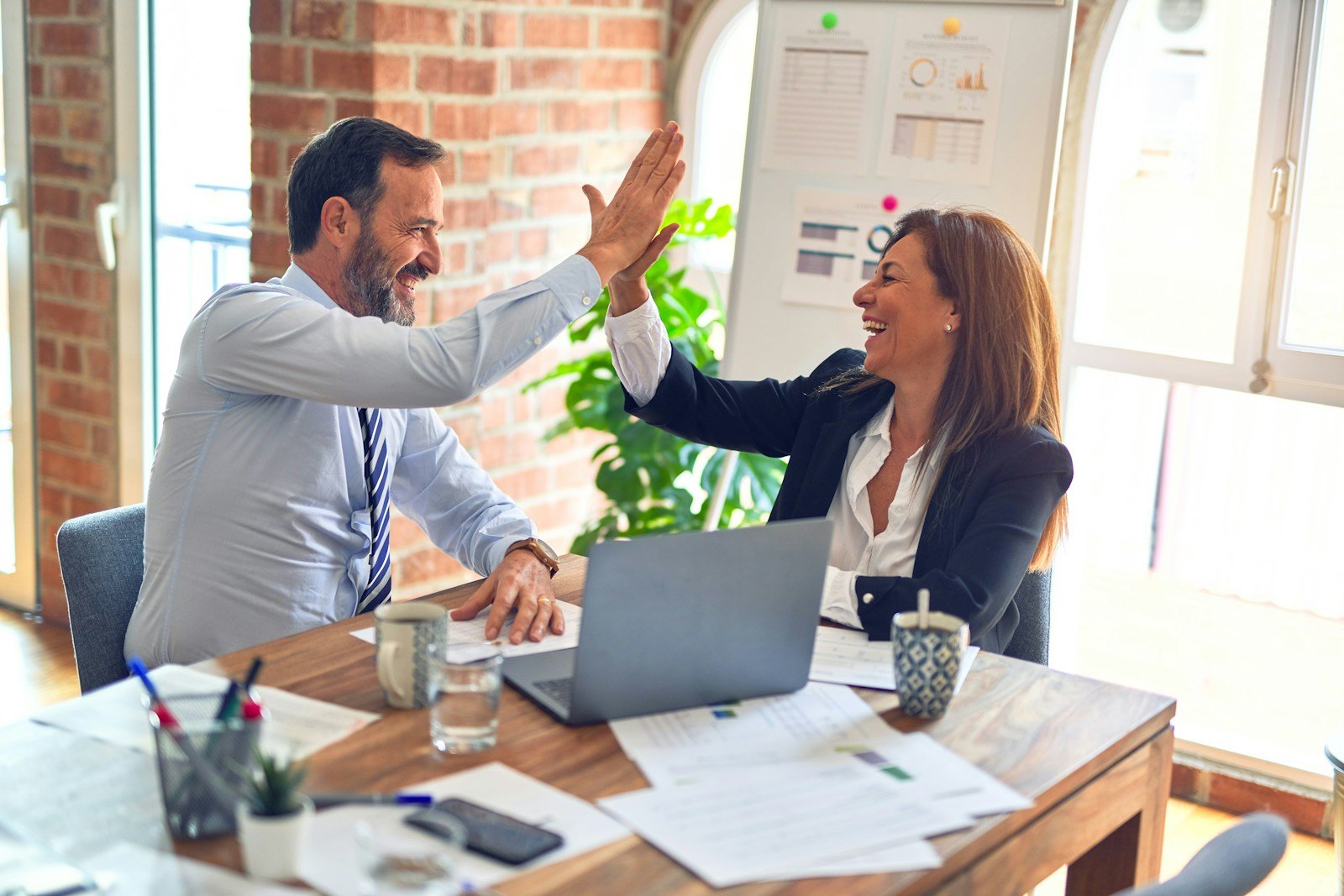 Two business professionals high-fiving at a desk with charts and data on a whiteboard behind them